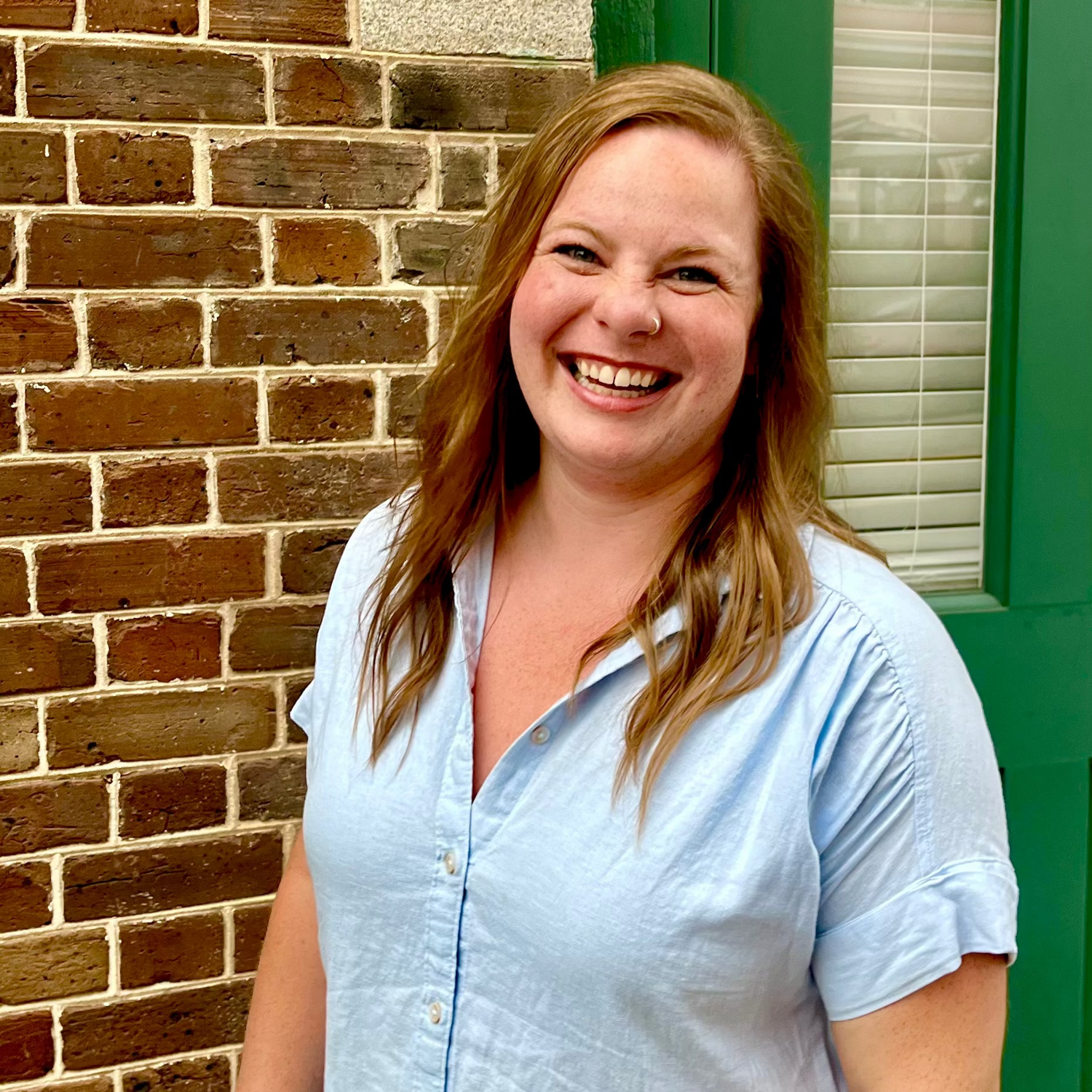 woman smiling in blue chambray dress in front of a brick wall and green door